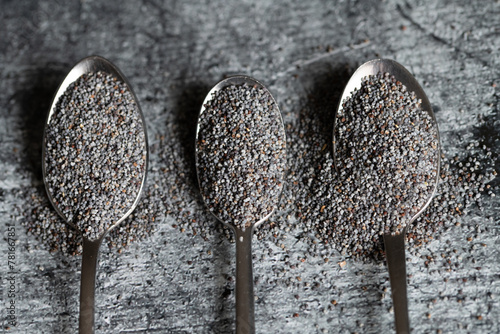 Close-up of three metal spoons lying next to each other against a gray background. There are unground poppy seeds on each of the three spoons. The poppy seeds can be used for cooking.