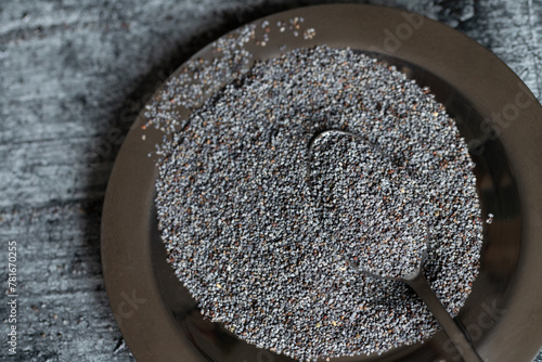 Close-up of a metal bowl containing blue poppy seeds. A spoon is in the poppy seed. The background is gray.