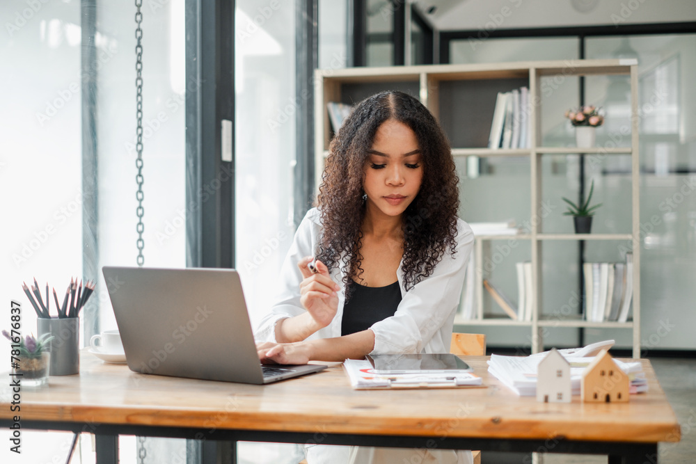 Real estate agent examines property documents while working on a laptop in a modern office, with miniature house models on the table. Real estate agent examines property documents while working on a laptop in a modern office, with miniature house models on the table.