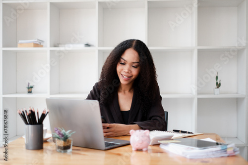 Cheerful financial advisor organizes client portfolios on her laptop, in a modern office with a piggy bank signifying savings on the desk.