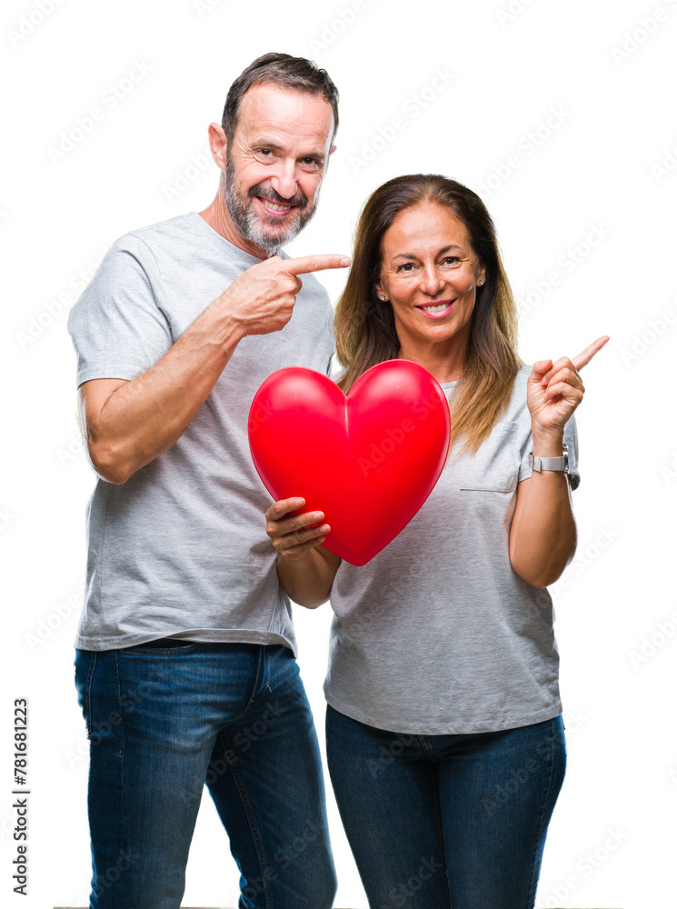 Middle age hispanic casual couple in love holding red heart over isolated background very happy pointing with hand and finger to the side