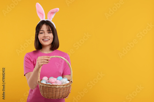 Easter celebration. Happy woman with bunny ears and wicker basket full of painted eggs on orange background, space for text
