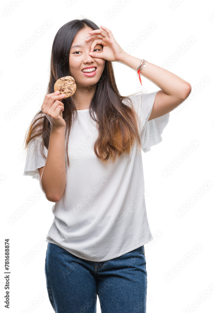 Young asian woman eating chocolate chip cookie over isolated background with happy face smiling doing ok sign with hand on eye looking through fingers Young asian woman eating chocolate chip cookie over isolated background with happy face smiling doing ok sign with hand on eye looking through fingers