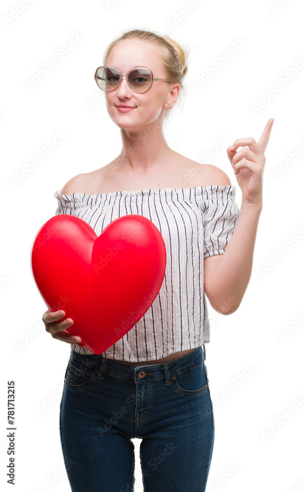 Blonde teenager woman holding red heart surprised with an idea or question pointing finger with happy face, number one Blonde teenager woman holding red heart surprised with an idea or question pointing finger with happy face, number one
