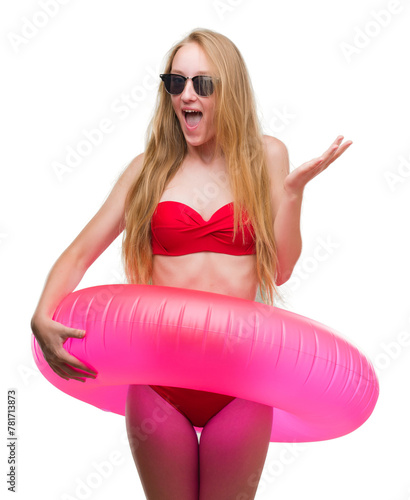 Blonde teenager woman wearing bikini and holding pink floater very happy and excited, winner expression celebrating victory screaming with big smile and raised hands photo