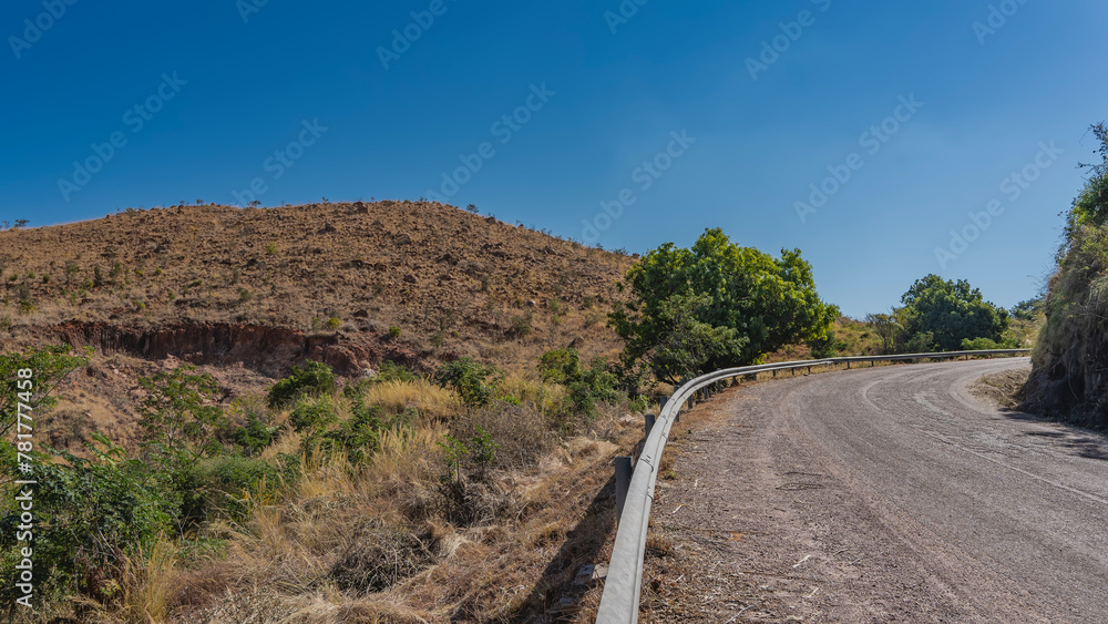 An asphalt road wraps around a hill. A protective barrier on the roadside. Green vegetation on red soil, Clear blue sky. Madagascar landscape
