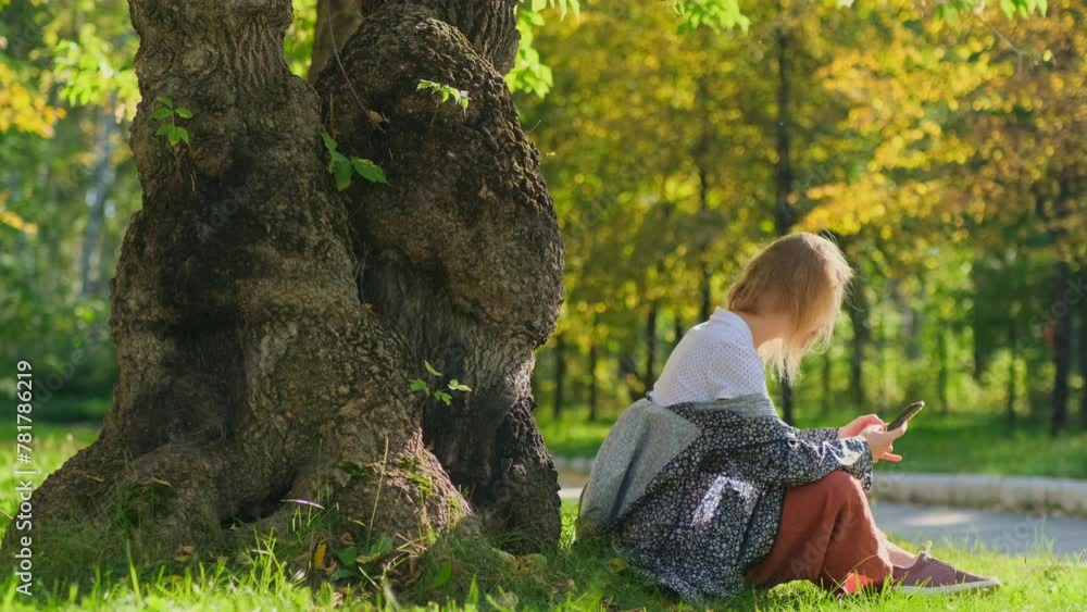 A pretty caucasian woman using her smartphone while sitting near big tree in an autumn park on a sunny day. A happy blonde is enjoying weekend outdoors. A pretty caucasian woman using her smartphone while sitting near big tree in an autumn park on a sunny day. A happy blonde is enjoying weekend outdoors.