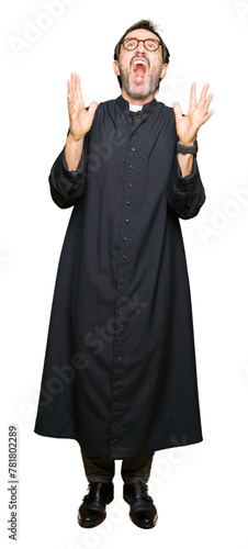 Middle age priest man wearing catholic robe crazy and mad shouting and yelling with aggressive expression and arms raised. Frustration concept.