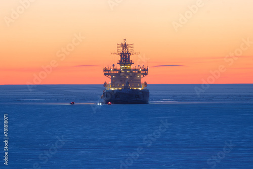 Icebreaker in the frozen sea. Beautiful winter arctic landscape. Icebreaker among the ice-covered sea expanses. Cars on the ice near a ship anchored in the roadstead. Evening twilight after sunset.