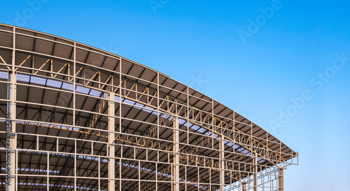 Part of large metal industrial factory building structure with corrugated steel curve roof and skylights in construction site against blue sky background, low angle view with copy space © Prapat