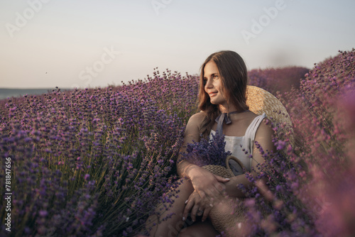 A woman is sitting in a field of lavender flowers. She is wearing a straw hat and holding a basket of flowers. The scene is peaceful and serene, with the woman enjoying the beauty of the flowers.