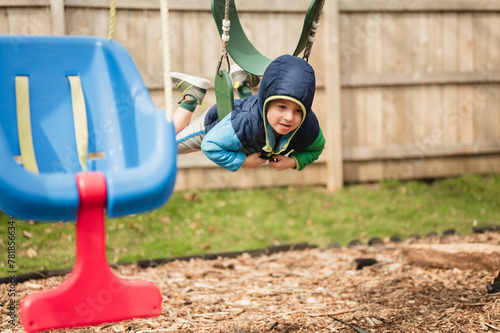Fotografie Happy child swinging on belly on swing in coat on playground
