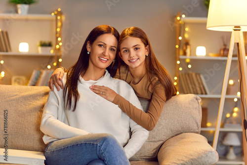 Portrait of smiling happy young mother with teen daughter sitting on sofa at home and looking cheerful at camera. Joyful woman with child girl hugging indoors. Mothers day, love and care concept.