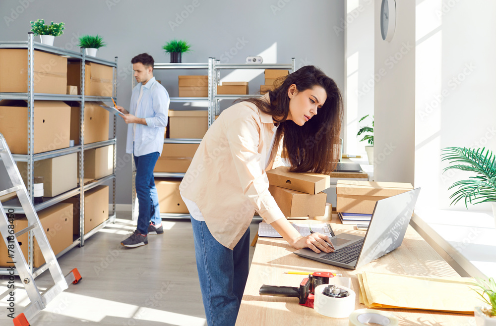 Female manager working on laptop at the desk in logistics or distribution center with male colleague preparing order for client in storeroom in warehouse. Inventory and delivery concept.