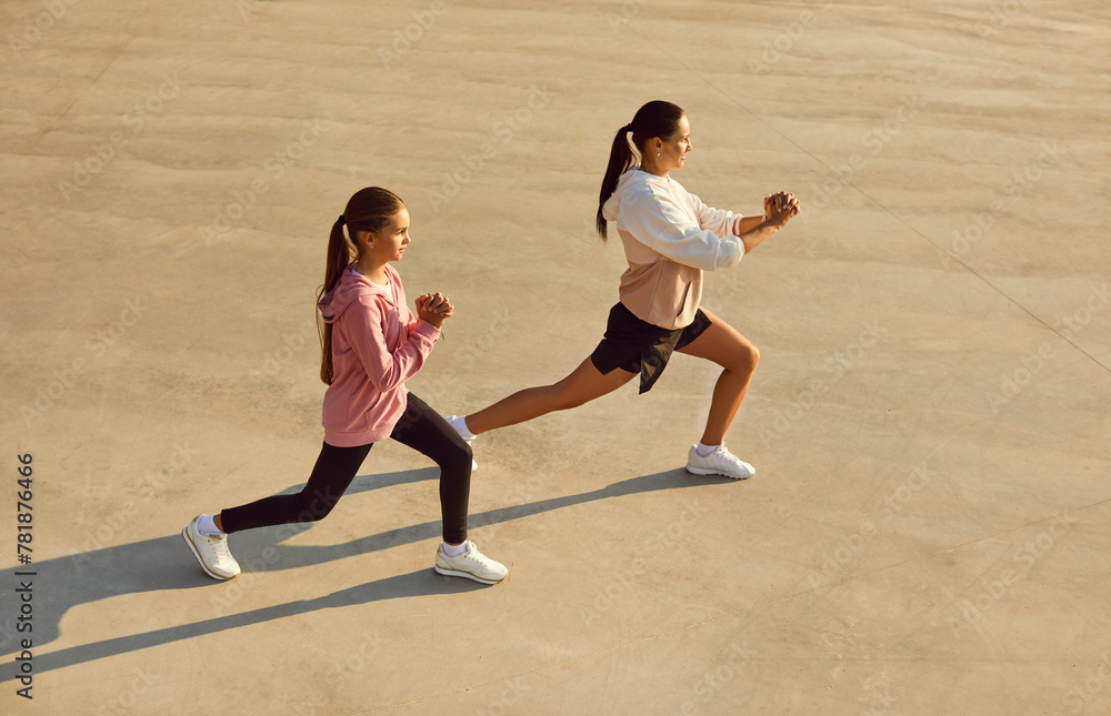 Mother and child having outdoor workout on sunny summer evening. Sporty confident strong woman and daughter in sportswear standing on concrete city sports ground and doing fitness exercises together