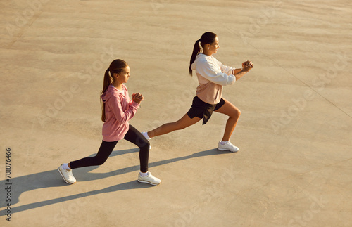 Mother and child having outdoor workout on sunny summer evening. Sporty confident strong woman and daughter in sportswear standing on concrete city sports ground and doing fitness exercises together
