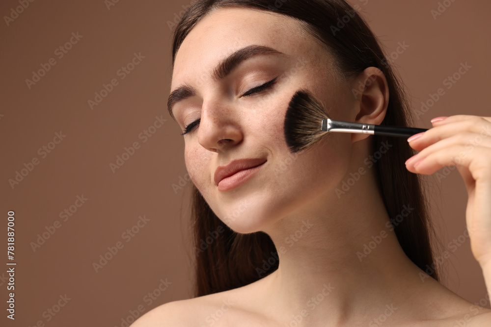 Beautiful woman with freckles applying makeup with brush on brown background, closeup
