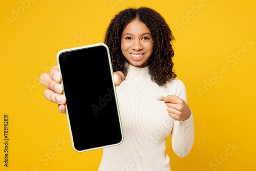 Little fun kid teen girl of African American ethnicity wear white casual clothes hold use point on blank screen area mobile cell phone isolated on plain yellow background. Childhood lifestyle concept.