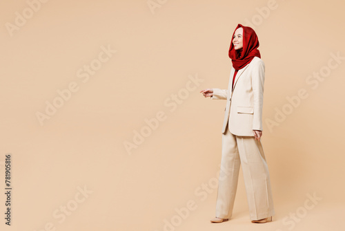 Full body side view smiling happy young Arabian Asian Muslim woman wearing red abaya hijab suit clothes walk go look aside isolated on plain beige background studio. UAE middle eastern Islam concept.