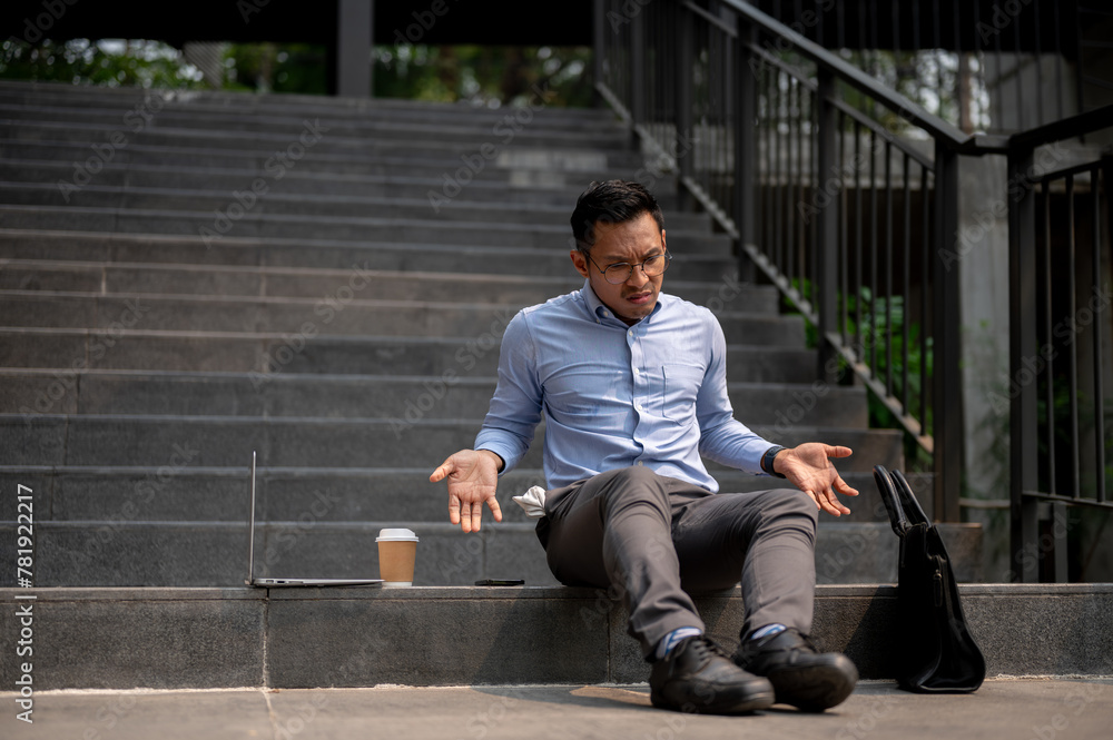An Asian businessman sits on outdoor steps with a laptop and a briefcase, expressing confusion.