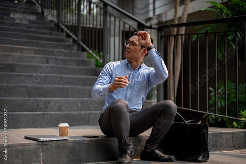 An Asian businessman sits on steps, looking upward with his hand shielding his eyes from the sun.