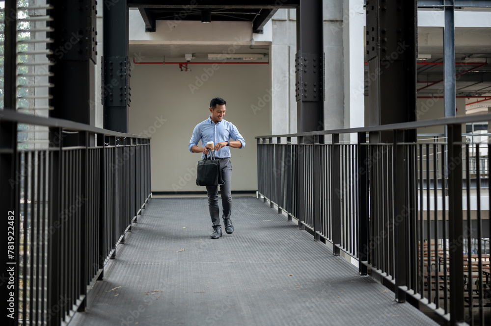 A confident Asian businessman is checking time on his wristwatch while walking along a corridor. A confident Asian businessman is checking time on his wristwatch while walking along a corridor.