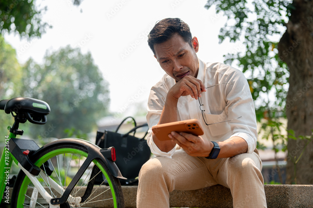 Sad Asian businessman sits outdoors, holding his phone with a concerned expression, deep in thought. Sad Asian businessman sits outdoors, holding his phone with a concerned expression, deep in thought.