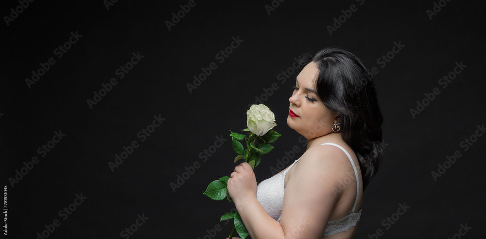A beautiful plump woman in lingerie with a rose in her hands poses in a dark studio. She is wearing a white bra and black tights.