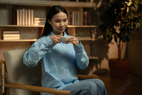 In the soft light of early morning, an Asian woman is seated comfortably in a home library setting, savoring the moment as she prepares a cup of tea, exuding calm and tranquility. 