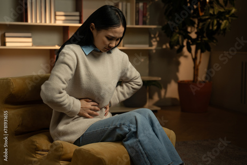 An Asian woman is clutching her stomach and appears to be in discomfort while seated on a couch in a cozy home library setting. The warm lighting suggests that it may be evening time.