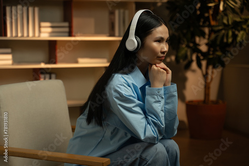 A pensive young Asian woman relaxes in a cozy living room setting, putting on her headphones to enjoy her favorite tunes as evening light filters through the room.