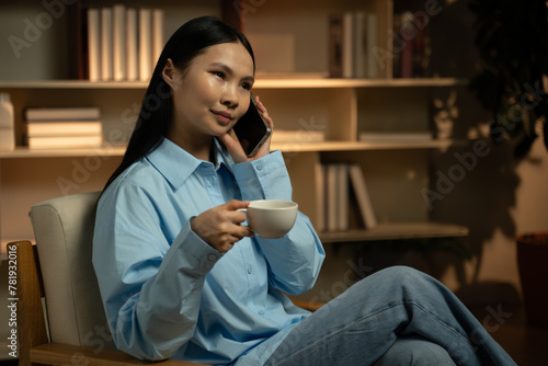 A young Asian woman is comfortably relaxing on a couch at home in the evening, enjoying a warm cup of coffee while engaged in a casual phone conversation. She appears content and at ease in her serene