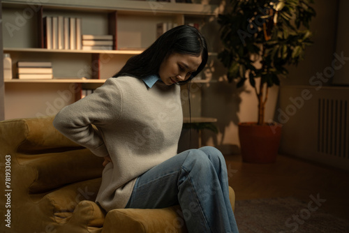 An Asian woman is seen sitting at the edge of a couch, clutching her lower back in discomfort, possibly indicating the onset of back pain.