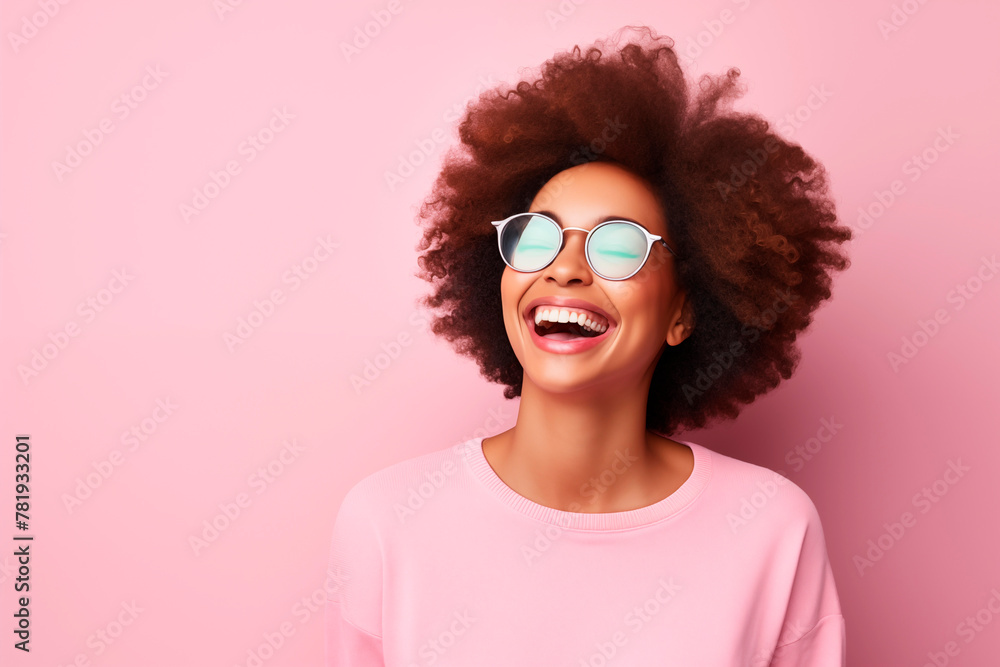 A joyful young woman laughing, wearing round sunglasses, set against a vibrant pink background; her afro hairstyle adds to her cheerful look.