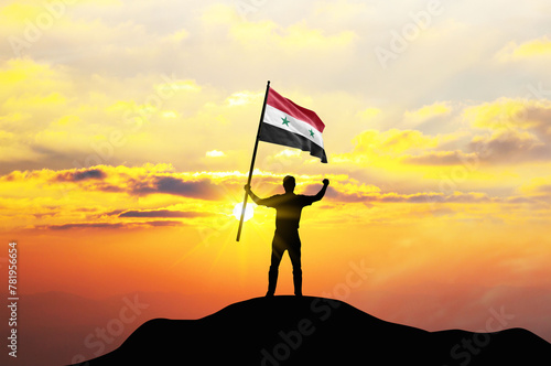 Syria flag being waved by a man celebrating success at the top of a mountain against sunset or sunrise. Syria flag for Independence Day.