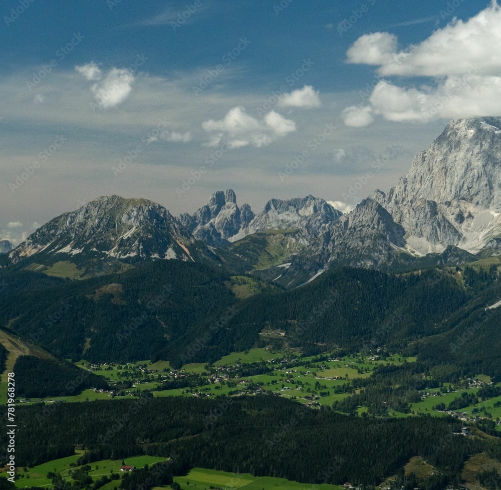 Fototapeta premium Aerial shot of a locality surrounded by trees and high rocky peaks covered in snow