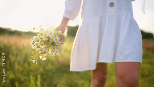 Woman in white dress and rubber boots walk along rural road with dirty muddy puddle enjoying nature, legs close-up. Female romantic holding bouquet of wildflowers in hands. Resting in countryside.