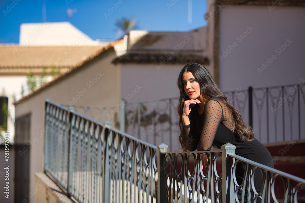 Young, beautiful, brunette woman in an elegant black dress, posing looking at camera leaning on a railing in a beautiful white Andalusian village. Concept beauty, fashion, trend, travel, rural. Young, beautiful, brunette woman in an elegant black dress, posing looking at camera leaning on a railing in a beautiful white Andalusian village. Concept beauty, fashion, trend, travel, rural.