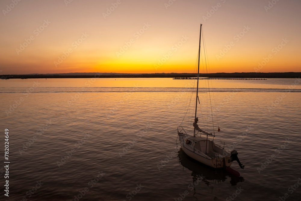 A pleasure sailboat anchored on a beach at sunset with orange sky on the horizon in the background