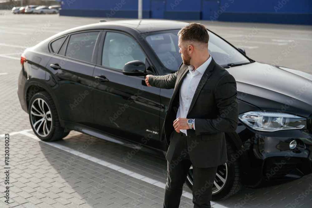 Locking the automobile. Businessman in suit is near his black car outdoors