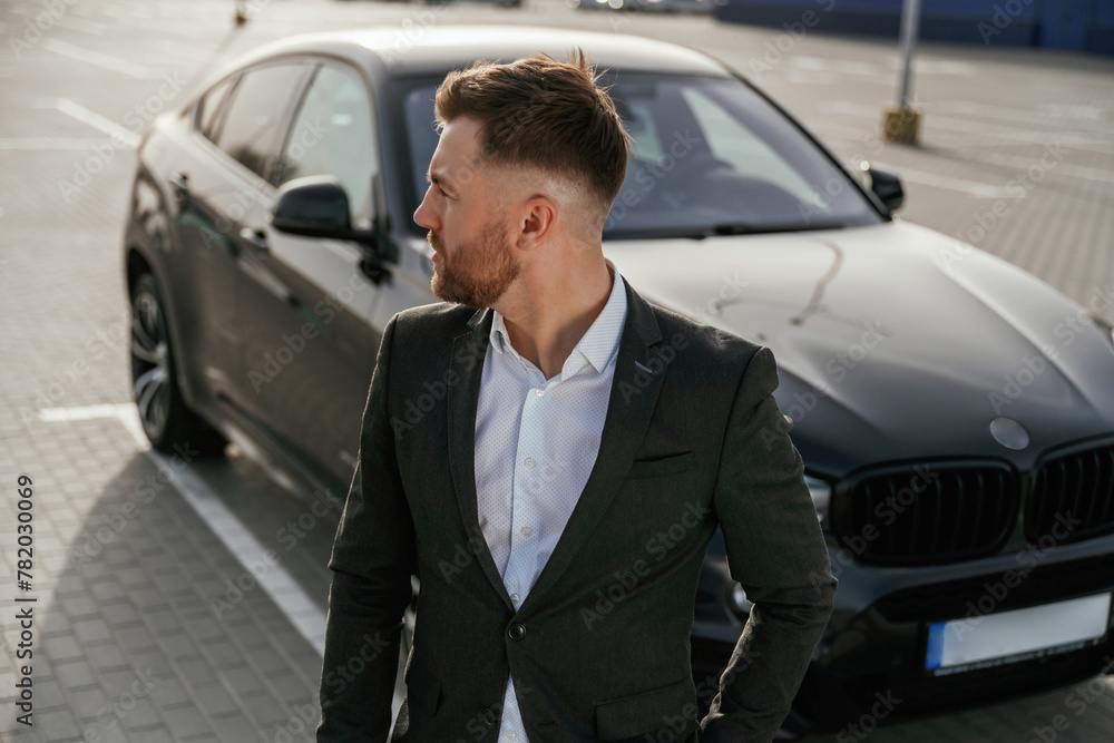 Focused portrait, against vehicle. Businessman in suit is near his black car outdoors Focused portrait, against vehicle. Businessman in suit is near his black car outdoors