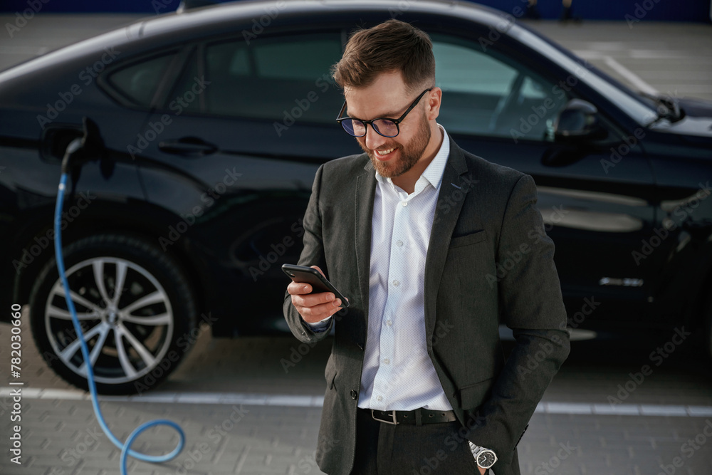 Smartphone in hand. Businessman in suit is near his black car outdoors