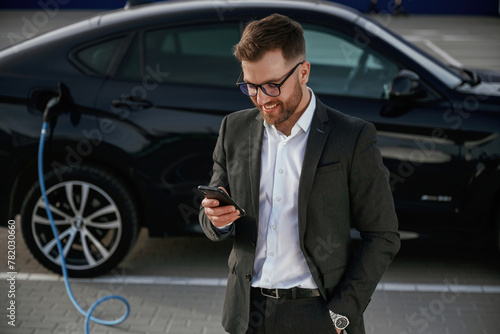 Smartphone in hand. Businessman in suit is near his black car outdoors © standret