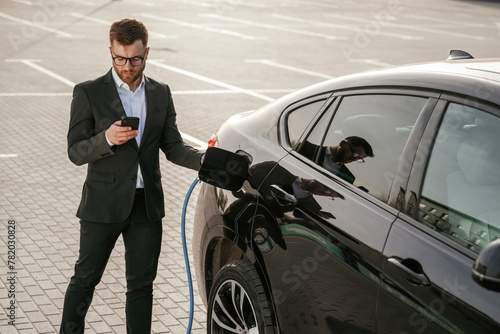 Holding phone in hand while vehicle is charging. Businessman in suit is near his black car outdoors © standret