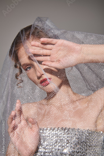 A young woman strikes a pose in a studio, dressed in a wedding gown with a veil gracefully flowing over her head.
