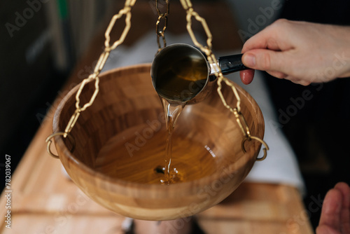 Close-up cropped shot of unrecognizable spa specialist pouring hot aromatic oil into bowl during preparation for exotic Shirodhara treatment, Ayurvedic healing technique in wellness center.