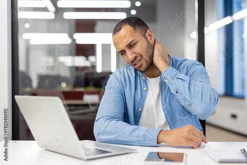 Tired hispanic man in shirt sitting in office at desk with laptop and holding hand on neck, doing massage, feeling pressure and pain