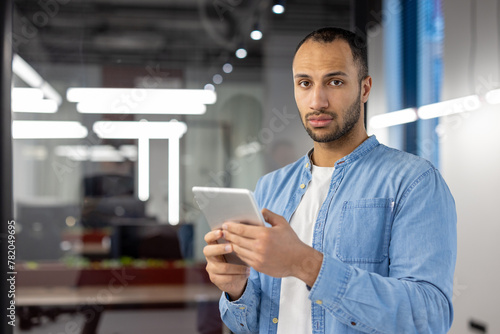 Portrait of a serious young Latin American man standing in the office, holding a tablet and looking at the camera