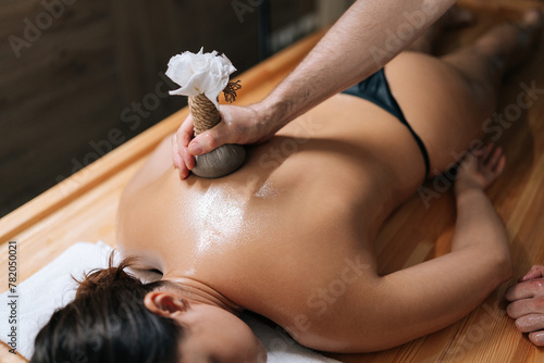 High-angle view of relaxed young woman lying with closed eyes receiving herbal bolus bags massage. Closeup hands of Ayurveda massage therapist pressing herbal bolus bags onto female body.
