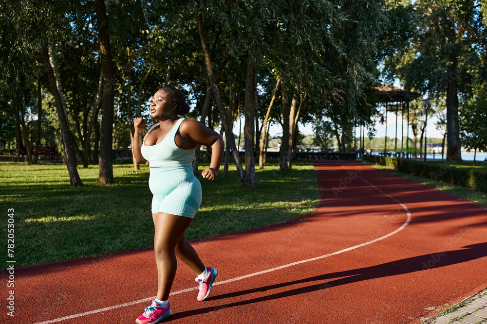 Obraz premium An African American woman in sportswear energetically runs along a track in a lush park setting, embodying positivity and confidence.